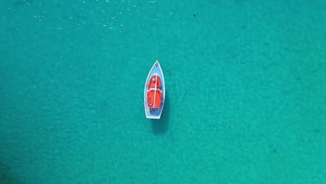 Orange-sailboat-with-blue-deck-bobs-in-shimmering-ocean-water,-aerial-static-top-down