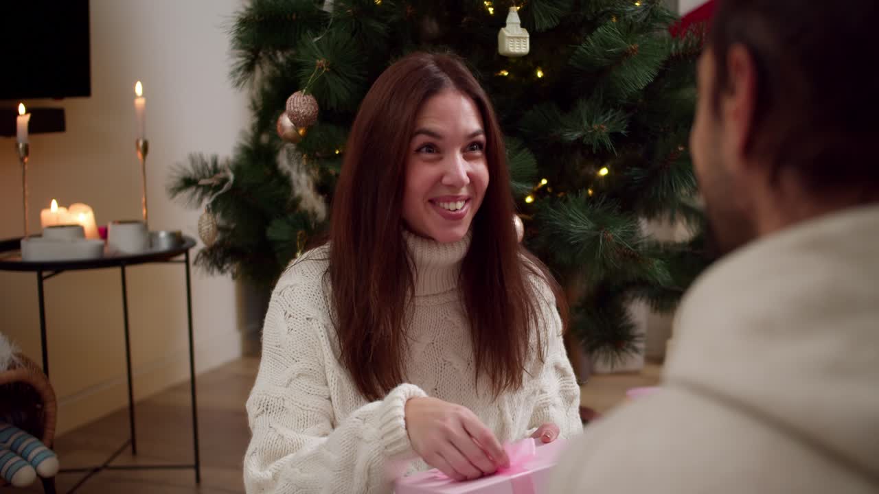 por encima del hombro, un tipo le da a su novia morena en un suéter de navidad blanco un regalo en una caja rosada con un lazo y la chica abre este regalo cerca del árbol de navidad por la noche en una casa acogedora