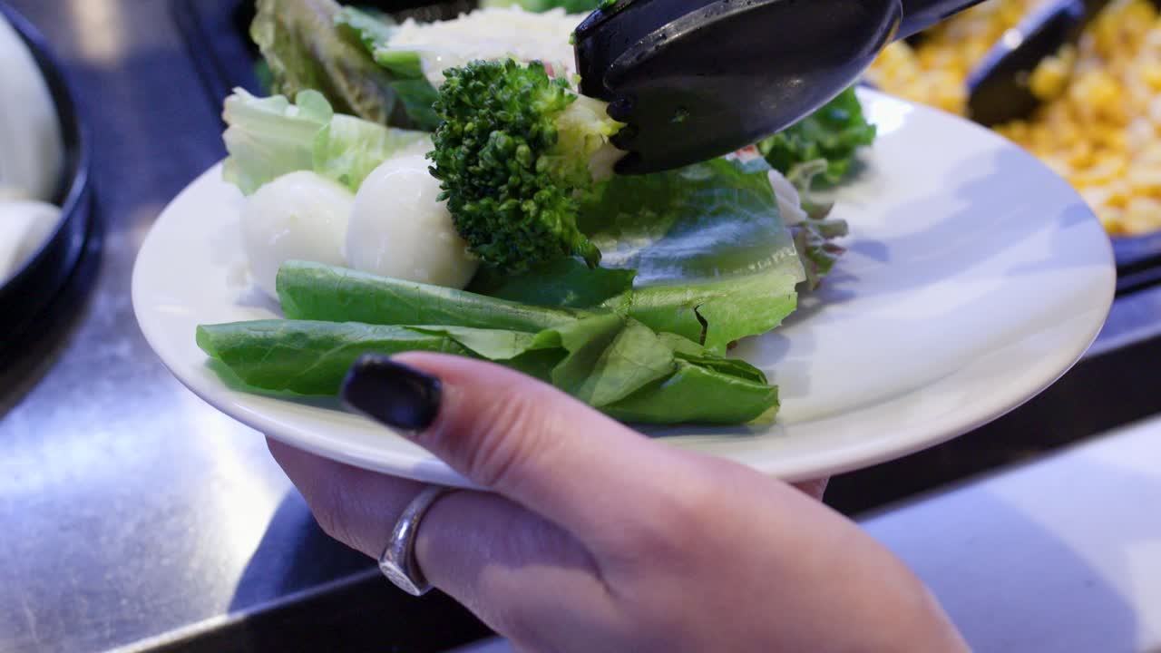 Close-up of woman filling plate with fresh salad ingredients at bright self-service buffet