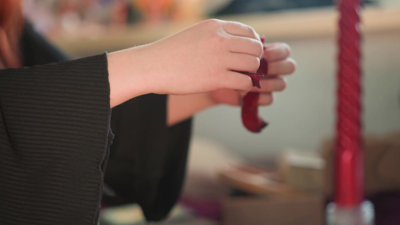 Closeup hands holding red wax pieces, performing wax reading ritual on wooden board with tall red candle, textured black sleeves, deliberate slow motions, warm candlelight and soft bokeh creating