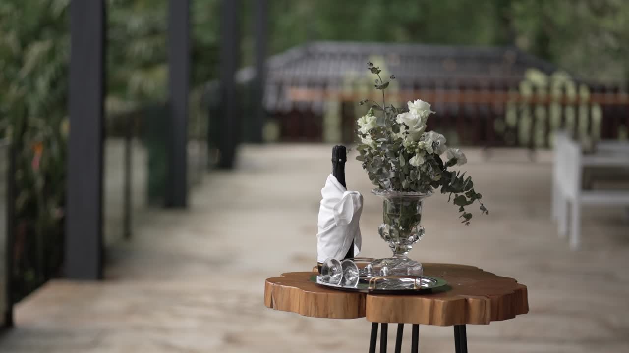 Small wooden table with floral centerpiece, wrapped wine bottle, and glassware in a garden setting
