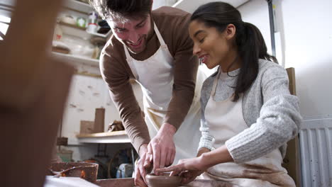 male teacher helping woman sitting at wheel in pottery class