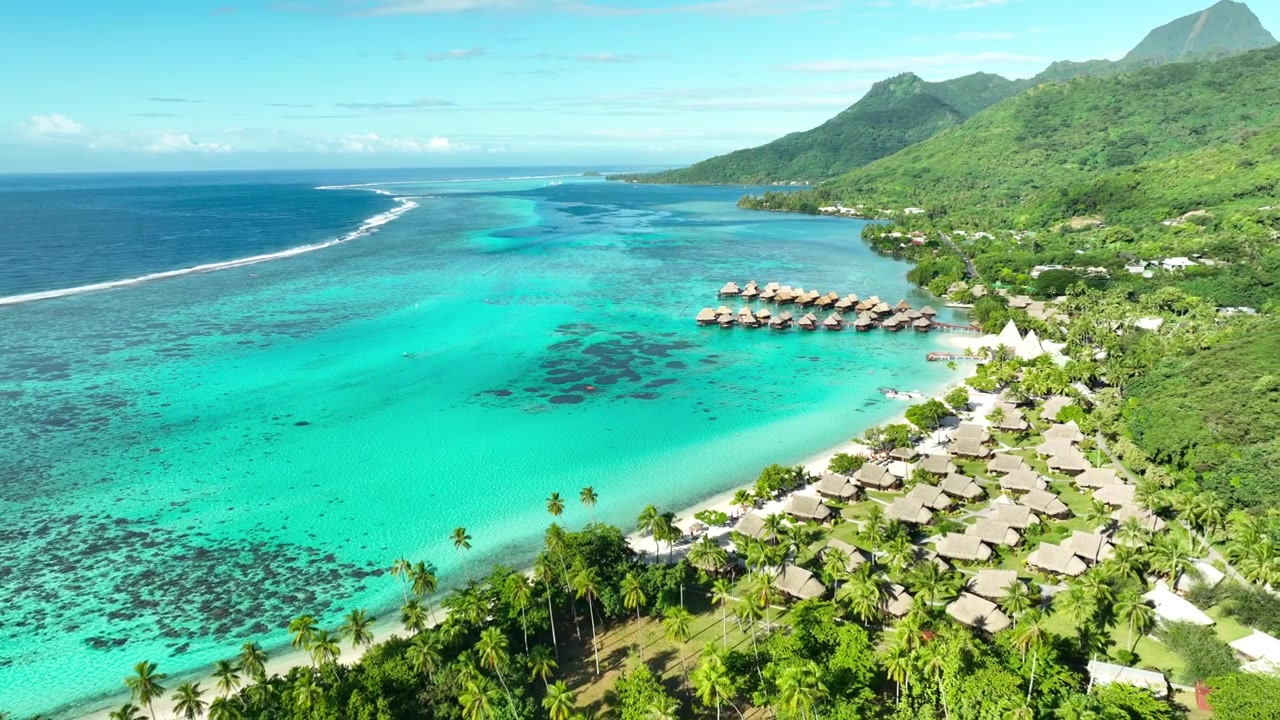Overwater bungalows stretch along reef edge beside turquoise lagoon at Temae Beach, Moorea, French Polynesia, dark coral patches and white sand line resort shoreline. Aerial drone backwards shot