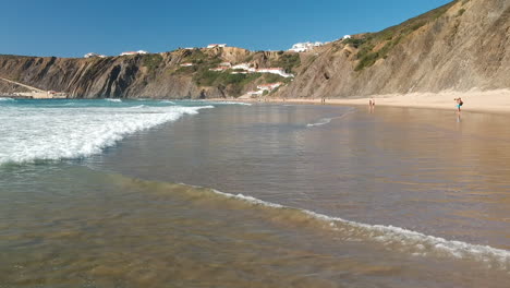 Toma-Aérea-De-Drones-De-Olas-De-Aguas-Bravas-Rodando-En-Una-Playa-Al-Lado-De-Algunas-Casas-En-Un-Acantilado-En-Algarve,-Portugal