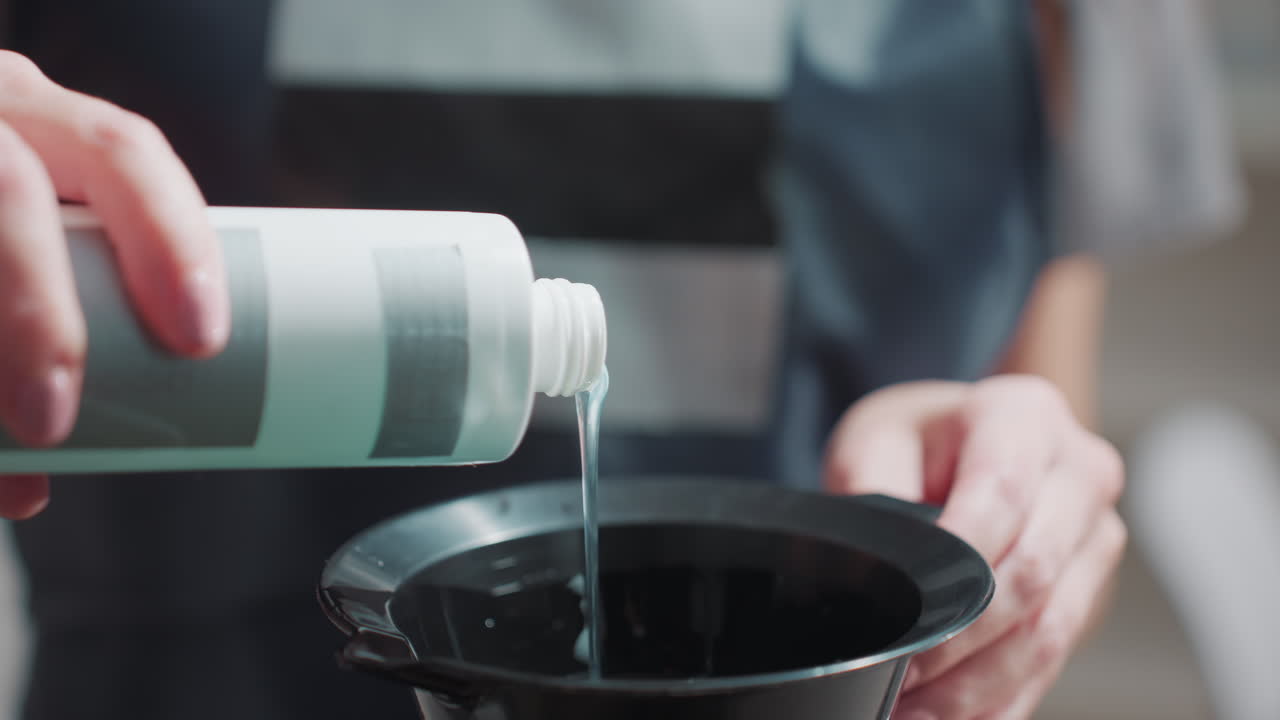 클로즈업 샷, 염색약을 그릇에 붓는 손, 계량된 액체, 검은색 혼합 용기, 부드러운 보케 배경, 앞치마 소매와 안정적인 손, 컬러 포뮬러를 위한 전문가의 준비, 은은한 살롱 톤