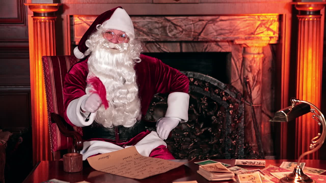 Santa Claus sitting indoors in front of fire place. Santa writing a letter with a red feather. Christmas cards beside the letter.