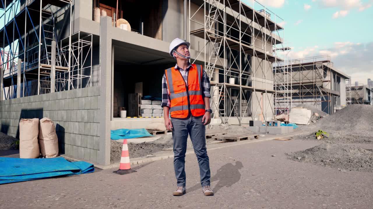 Full Body Of Asian Male Engineer With Safety Helmet Looking Around While Standing At Building & Construction Site in Progress to New House