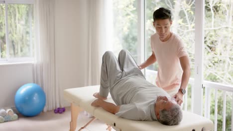 diverse male physiotherapist advising and senior male patient exercising on couch, in slow motion