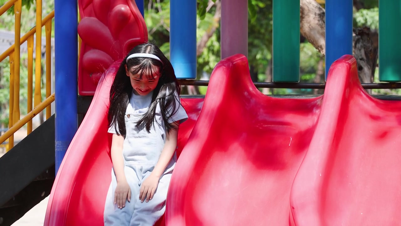 Una niña asiática alegre con peto vaquero saluda y se desliza por un tobogán rojo brillante