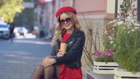 young beautiful woman in a red beret and dress drinking coffee and looks at the camera. photo shoot on the street