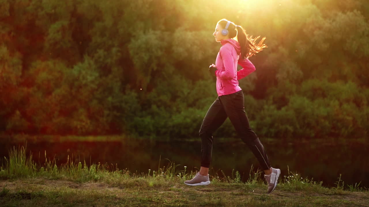 A girl in a pink jacket and black pants runs near the river in headphones preparing for the marathon