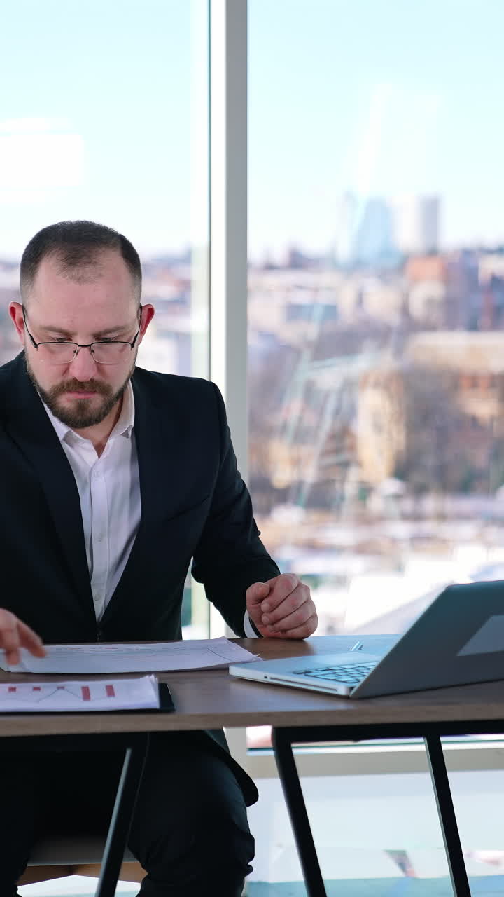 Busy man in suit working in office. Middle-aged entrepreneur in glasses sitting at table with many papers and a laptop and working seriously. Vertical video