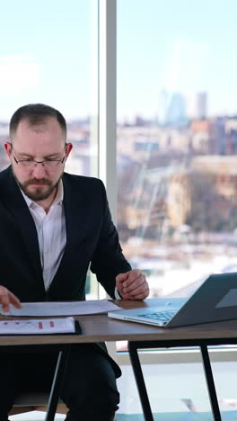 busy man in suit working in office. middle-aged entrepreneur in glasses sitting at table with many papers and a laptop and working seriously. vertical video