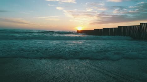 Toma-Aérea-De-La-Puesta-De-Sol-En-La-Playa-De-Tijuana-Con-El-Muro-Fronterizo