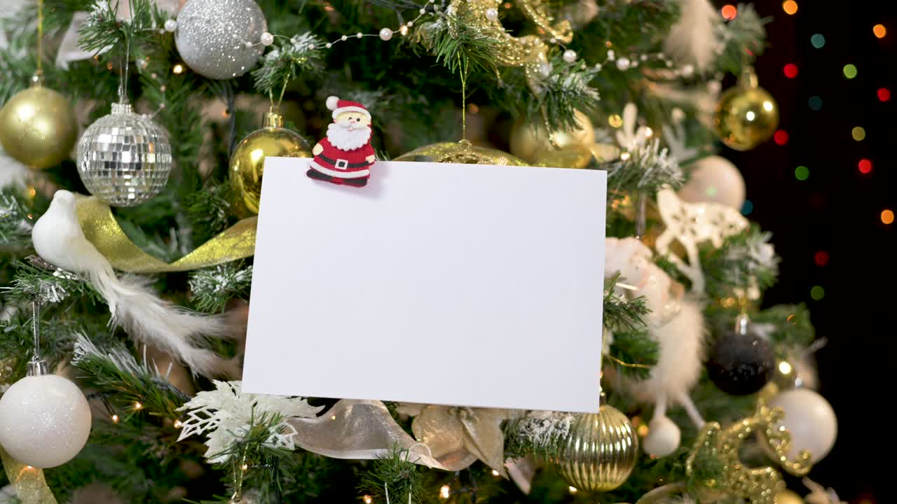 papel blanco para tarjetas de felicitación enganchado con ornamento de santa en oro árbol de navidad decorado en blanco con luces parpadeantes