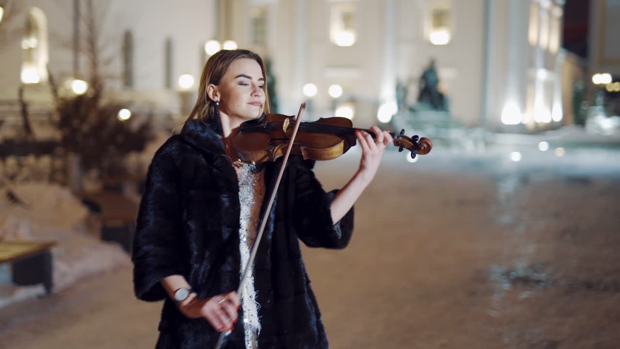 A young girl in a black coat is playing the violin on the background of buildings and evening lights in the centre of the city in the winter. Blurred Background
