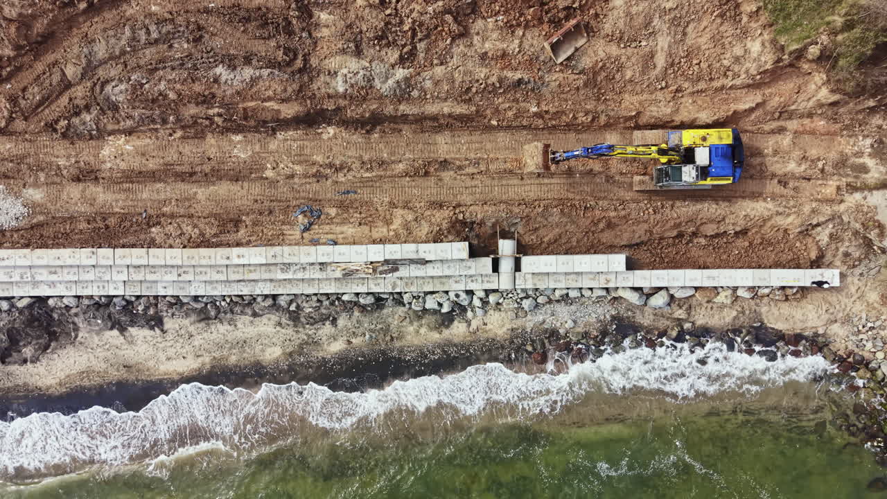 Costruzione di una struttura in cemento lungo la riva con macchinari pesanti che lavorano a terra vicino all'acqua durante una giornata nuvolosa.