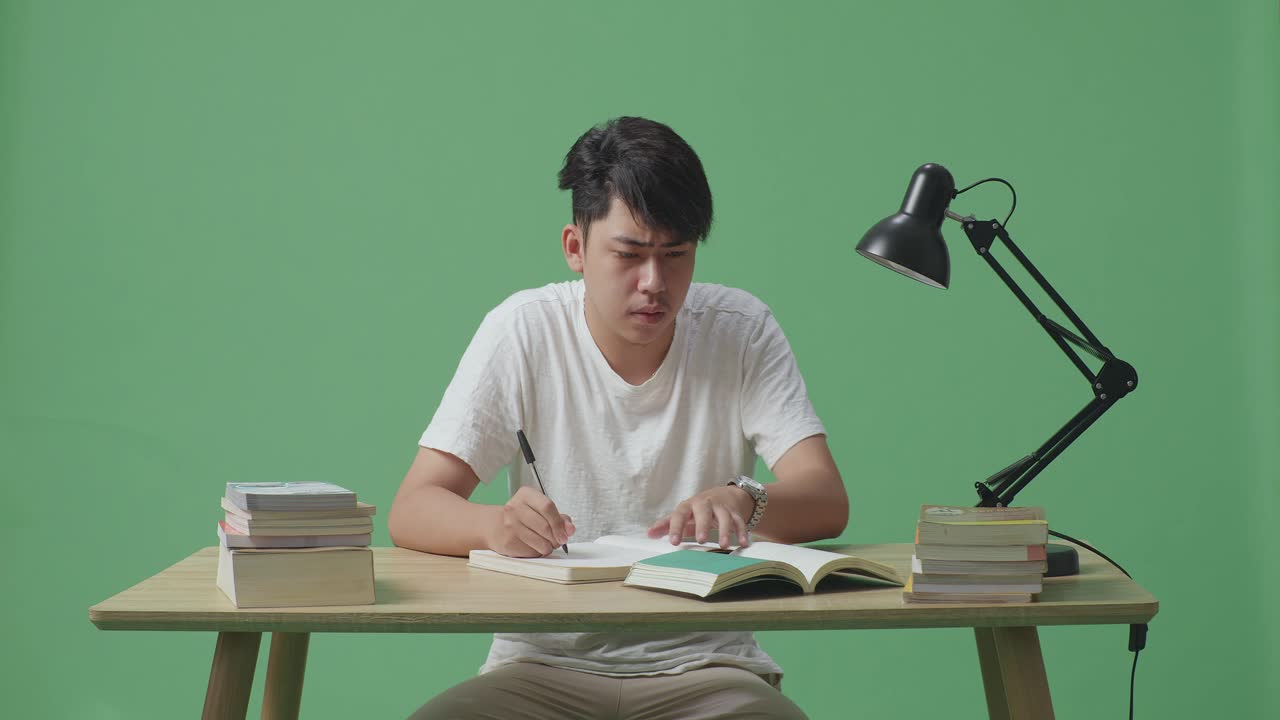 젊은 아시아 남성이 뭔가에 대해 생각하고 집에서 녹색 화면 배경에 책에 쓰는 동안 주위를 둘러보고 있습니다.