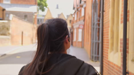 Rear-View-Of-Young-Woman-Exercising-Running-Along-Urban-Street-Wearing-Wireless-Earbuds