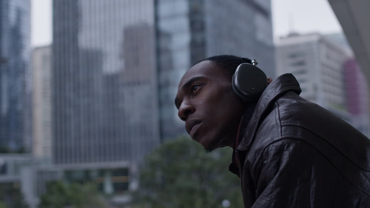 Modello afroamericano che guarda seriamente il paesaggio urbano di una smart city, mentre ascolta musica, indossando cuffie eleganti.