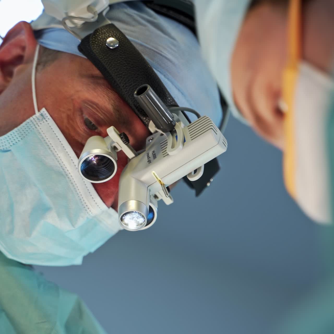 Adult male doctor wearing device binoculars at operation. Portrait of a surgeon in mask talking. Low angle view