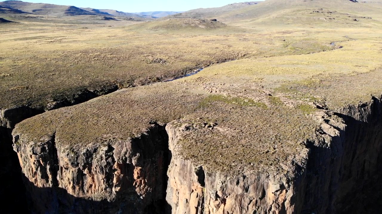 Vista aerea di un altopiano erboso sulla scogliera con un ruscello serpeggiante, toni dorati tenui, vegetazione bassa e rada, roccia verticale a strapiombo. Media