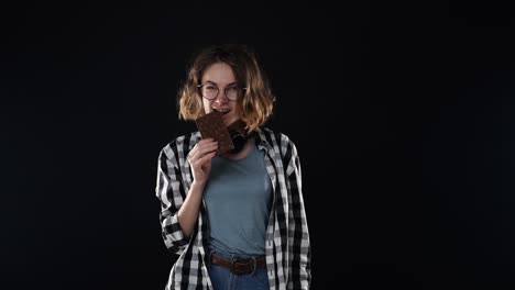 Sonriente-Hermosa-Joven-Morena-Con-Camisa-A-Cuadros-Y-Jeans-Posando-Comer-Barra-De-Chocolate-En-La-Mano-Con-Nueces-Aisladas-Sobre-Fondo-Negro-En-El-Estudio.-La-Gente-Emociones-Sinceras-Concepto-De-Estilo-De-Vida.