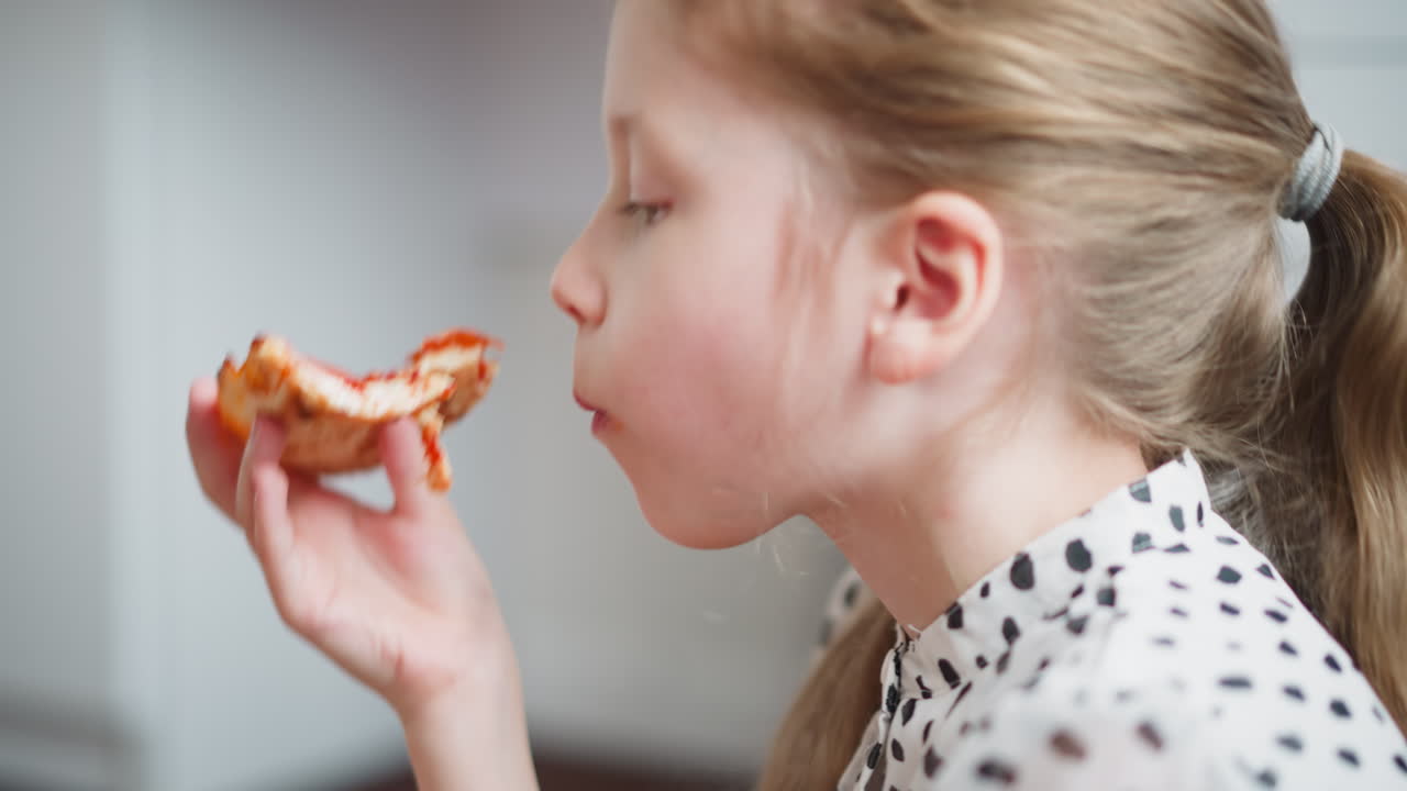 Side view of young girl with hair tied back in ponytail wearing spotted clothes enjoying slice of pizza indoors, biting eagerly into cheesy pepperoni topping with joyful expression