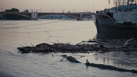debris floating in the river next to a docked boat in budapest, bridge visible in the background
