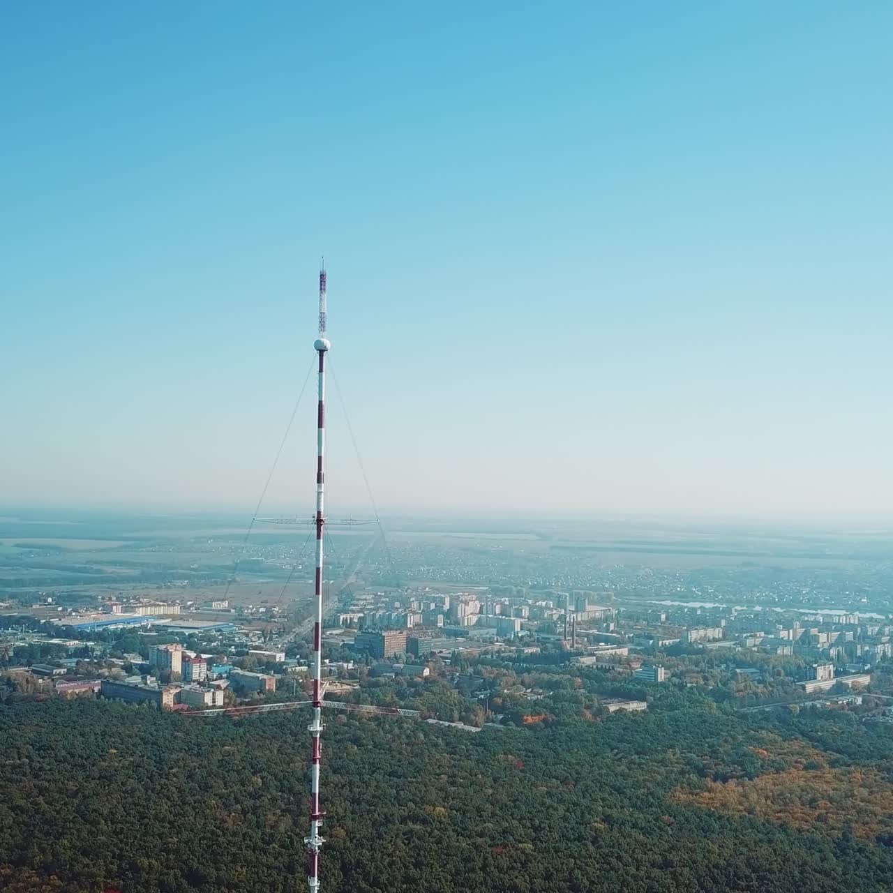 top of the telecommunication tower in red and white colours on the background of the cityscape and blue sky. Camera motion up