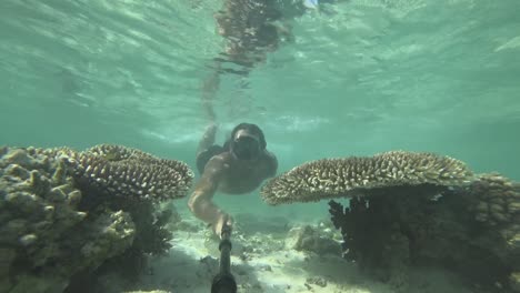 A-young,-fit-and-strong-man-with-long-hair-and-beard-is-swimming-in-very-shallow-water-next-to-coral-reef-and-white-sand-with-reflections-dancing-on-the-surface-above-him-in-vibrant-water