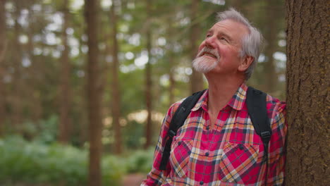 active retired senior man walking in woodland countryside wearing backpack leaning  on tree