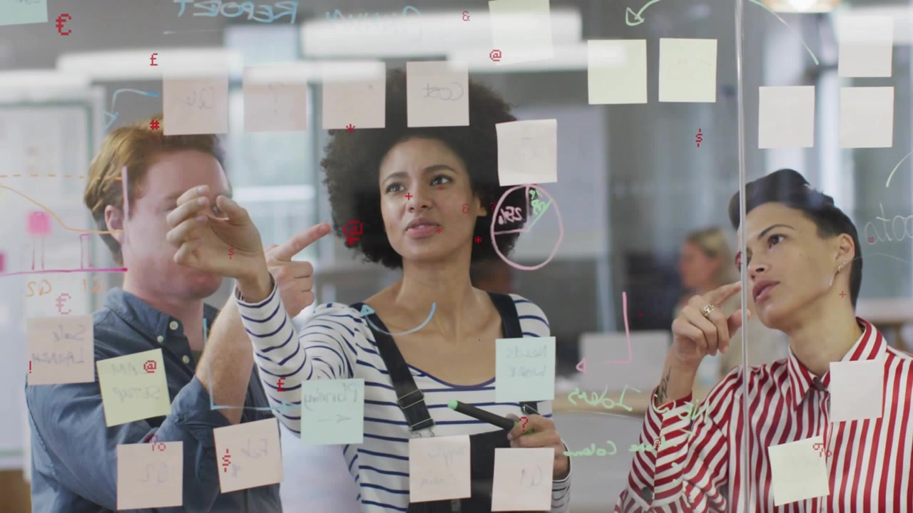 3 coworkers clustering at glass wall, center reaching up moving sticky notes for product plan