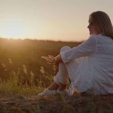 Eine-Frau-In-Einem-Weißen-Anzug-Sitzt-Auf-Einem-Hügel-Und-Bewundert-Den-Sonnenuntergang