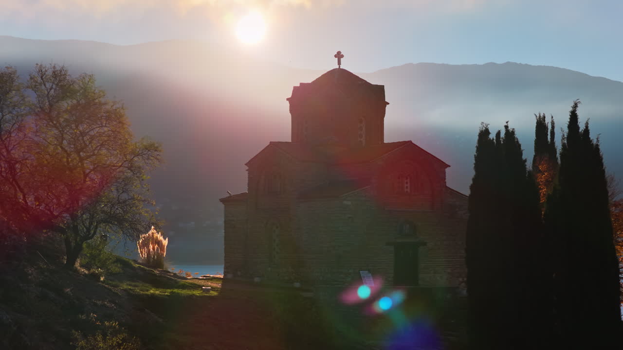 Aerial drone view of a sunrise scene behind St. John at Kaneo, with rays breaking through the clouds and illuminating the church's silhouette. North Macedonia