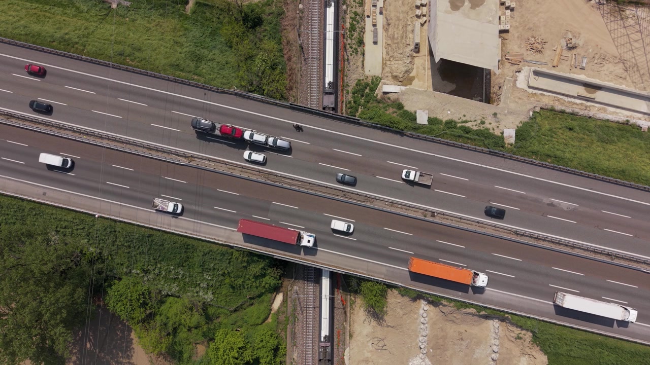 Veduta aerea di un treno che passa sotto un ponte autostradale nell'Italia settentrionale. Il traffico ferroviario e stradale simultaneo mostra una densa infrastruttura di trasporto e un sistema di mobilità.