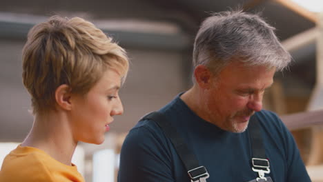 female apprentice learning from mature male carpenter with laptop in furniture workshop