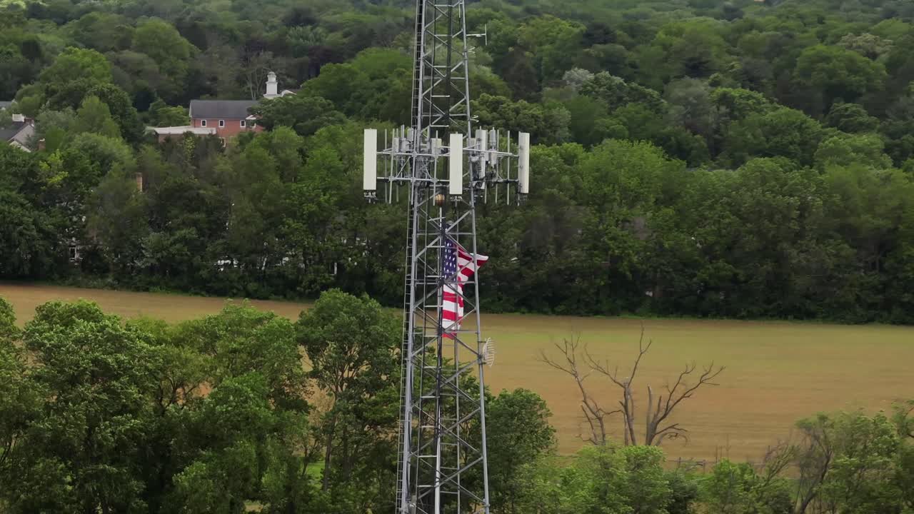BLowing american flag on flagpole beside transmission tower in rural area with green trees. Aerial close up shot. Cloudy day with Agricultural farm fields in distance.