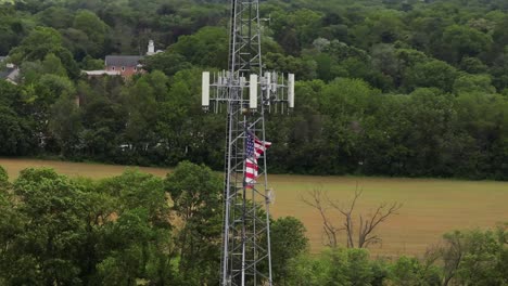 blowing american flag on flagpole beside transmission tower in rural area with green trees. aerial close up shot. cloudy day with agricultural farm fields in distance.