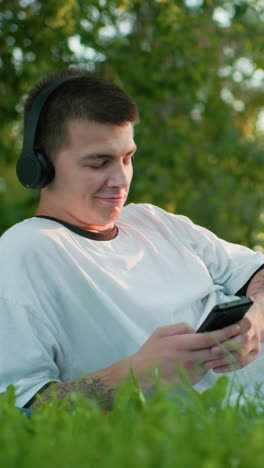 trabajador remoto acostado en un campo cubierto de hierba con auriculares, asintiendo con la cabeza a la música con una sonrisa sutil mientras opera el teléfono con la mano tatuada, rodeado de exuberante vegetación