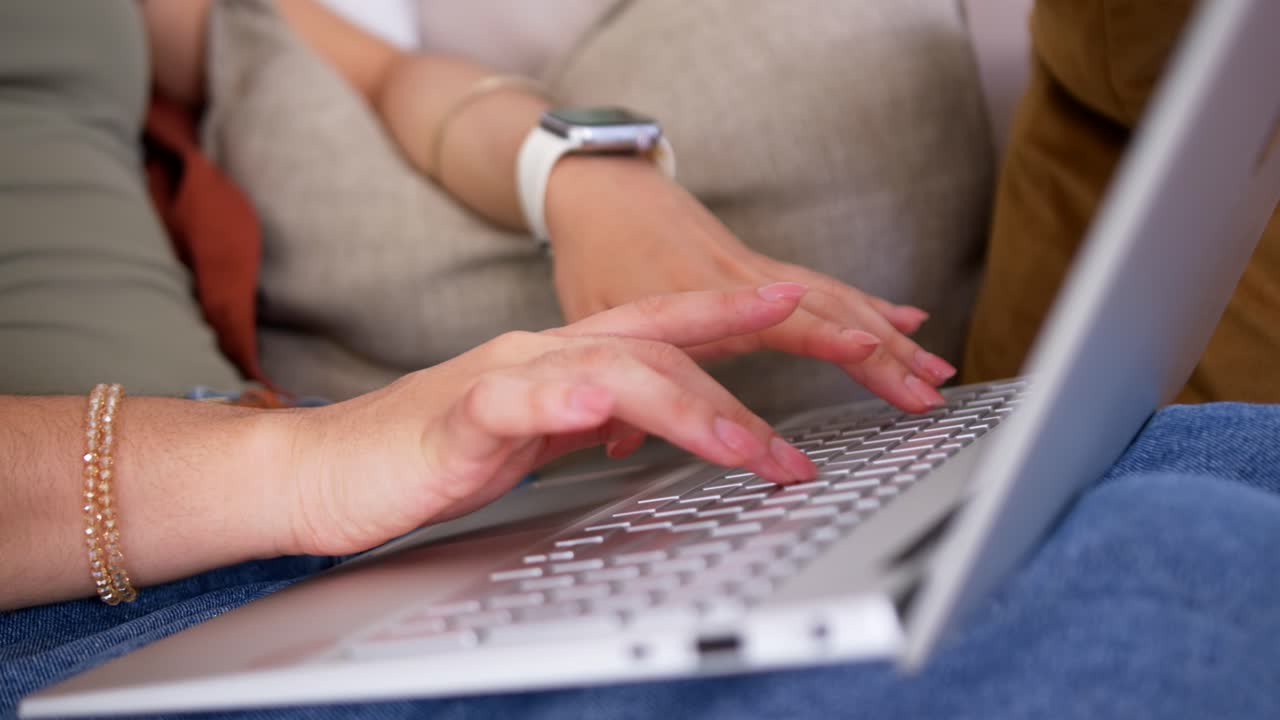 To veninder samarbejder, taster på en bærbar computer på sofaen, iført hvidt ur og guldbarmbånd.