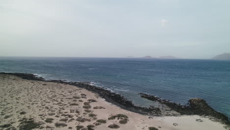 Playa-de-San-Juan,-Famara,-Lanzarote,-Canary-Island,-drone-over-the-sea-mountains-in-the-horizon
