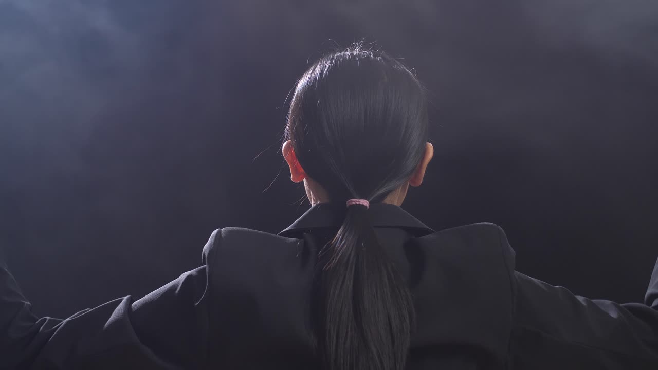 뒷면에서 가까이 쳐다보는 아시아의 지휘자 여성이 막대기를 들고 눈을 감고 안개와 함께 검은 스튜디오에서 몸짓을 보여줍니다.