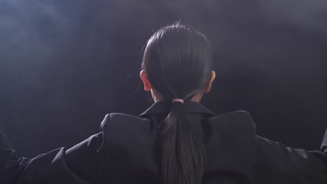 뒷면에서 가까이 쳐다보는 아시아의 지휘자 여성이 막대기를 들고 눈을 감고 안개와 함께 검은 스튜디오에서 몸짓을 보여줍니다.