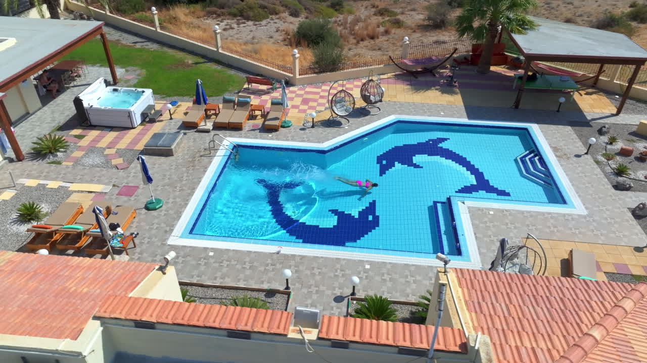 Aerial view of a woman jumping and swimming in a pool, at a villa, sunny day