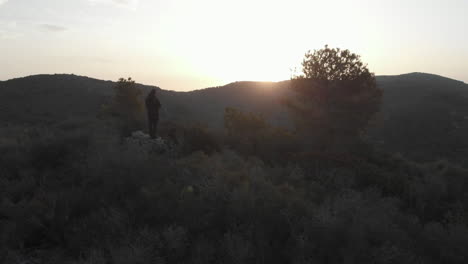 Drone-reveals-silhouette-of-man-hill-scape-during-sunset,-valley-and-bright-sky-contrast