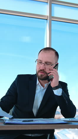 serious businessman working in office. bearded man boss in suit sitting at the desk with a laptop and talking the phone. business concept. vertical video
