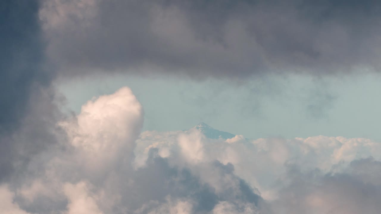 Distant view of Mount Teide covered in snow, rising above the clouds, seen from Gran Canaria on a moody winter day. A breathtaking contrast of volcanic peaks and dramatic sky.