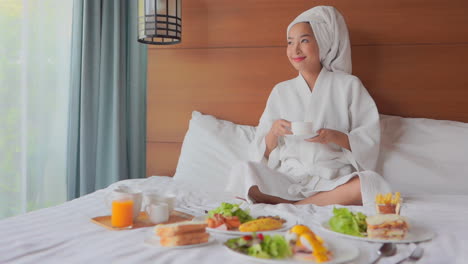 귀여운 아시아 젊은 여성이 침대에 앉아 흰색 목욕 가운을 입고 차를 마신다