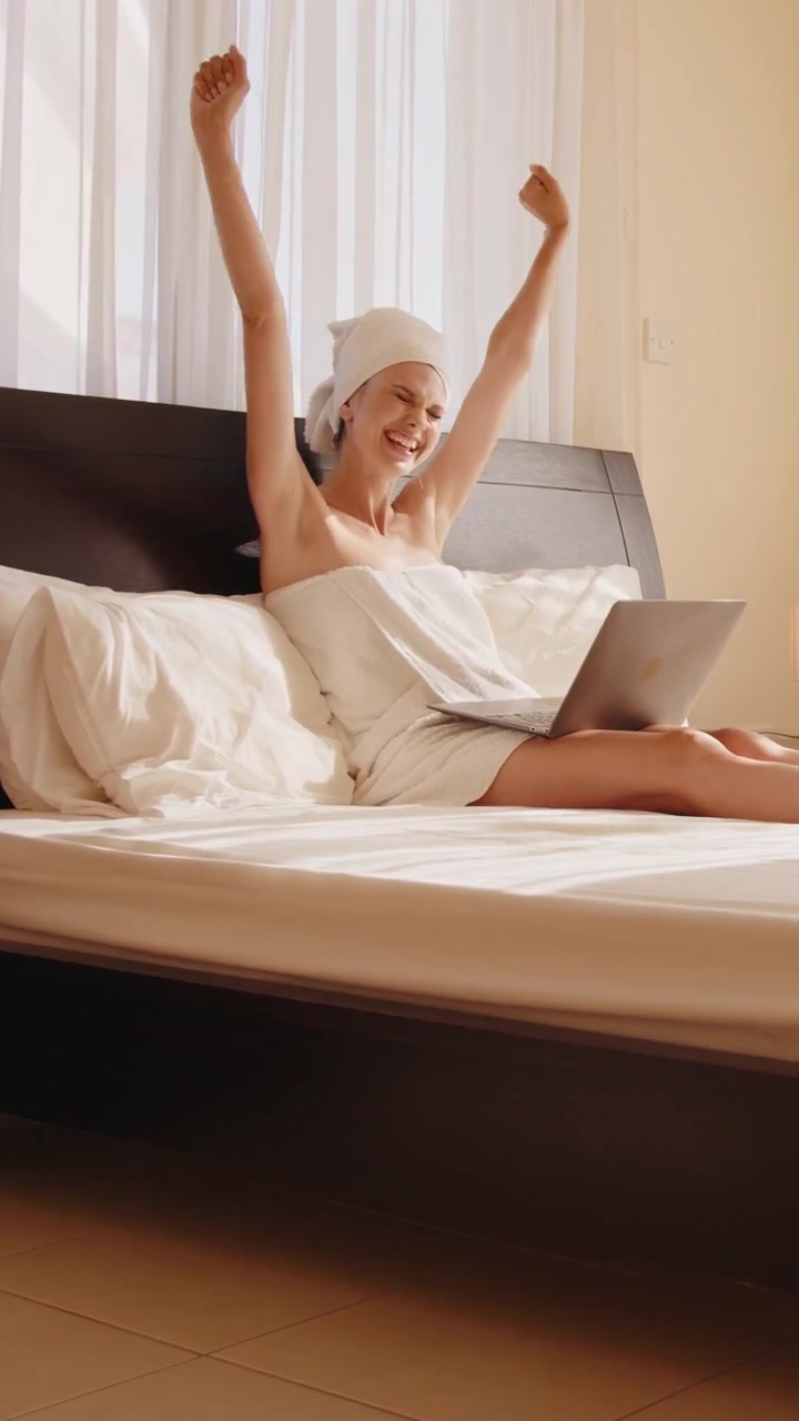 Una donna avvolta in un asciugamano sorride mentre usa un laptop su un letto in una stanza luminosa, video verticale.
